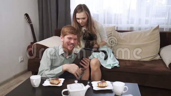 一对夫妇坐在家里的沙发上看着平板电脑微笑着喝着白色茶具上的茶看着视频的预览图