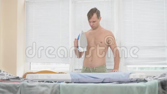 年轻男子裸露的躯干站在板后使用蒸汽熨斗熨烫衬衫在家务4k慢镜头视频的预览图