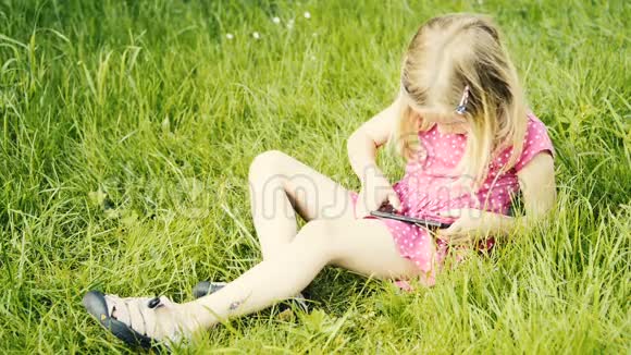 学龄前可爱的金发女孩坐在草地上玩智能手机视频的预览图