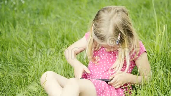 学龄前可爱的金发女孩坐在草地上玩智能手机视频的预览图