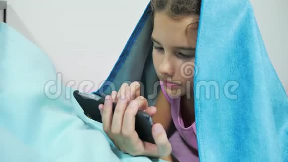 十几岁的女孩在毯子下玩智能手机在室内睡觉前的小女孩躺在床上上网视频的预览图
