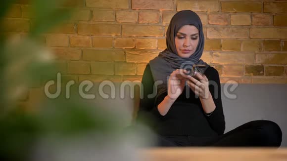 戴着头巾坐在室内沙发上用手机拍摄的穆斯林成年女性特写照片视频的预览图