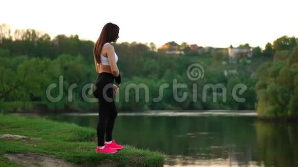 穿着白色上衣黑色紧身裤和粉色运动鞋的运动型黑发女郎在阳光下伸展肌肉准备视频的预览图