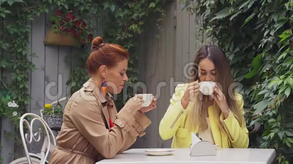 一个穿着棕色外套的中年红发女人和一个穿着黄色外套的棕色头发的年轻女人坐在一家夏季街头咖啡馆里视频的预览图