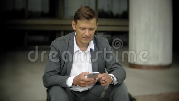商人坐在城市的楼梯上他穿着西装和公文包在智能手机上聊天视频的预览图