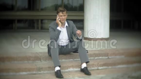 商人坐在城市的楼梯上他穿着西装和公文包在智能手机上聊天视频的预览图