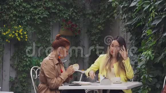 一个穿着棕色外套的中年红发女人和一个穿着黄色外套的棕色头发的年轻女人坐在一家夏季街头咖啡馆里视频的预览图