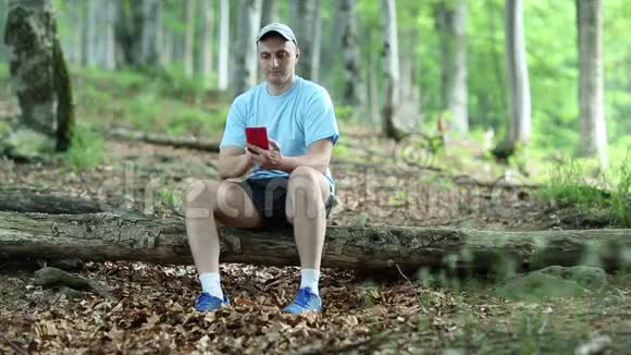 一个戴着红色智能手机的人坐在森林里的一棵倒下的树上视频的预览图