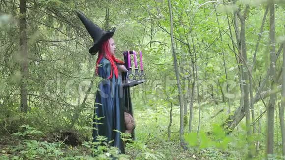 年轻的女巫红色的头发黑色的帽子和黑色的斗篷在森林里的蜡烛视频的预览图