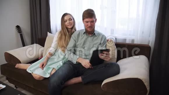 一对夫妇坐在家里的沙发上看着平板电脑微笑着喝着白色茶具上的茶看着视频的预览图