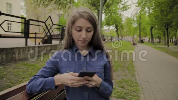 美丽的快乐女孩坐在城市公园的长凳上带着手机视频的预览图