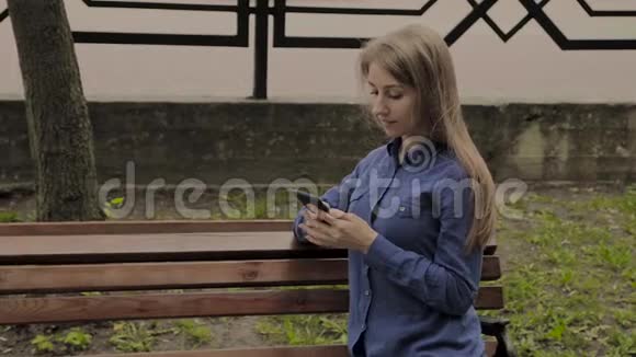美丽的快乐女孩坐在城市公园的长凳上带着手机视频的预览图
