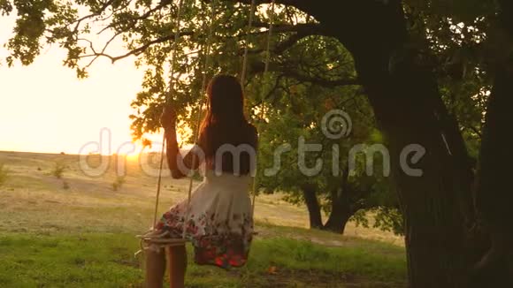 年轻女孩在橡木树枝上的绳索秋千上摆动公园里穿着白色裙子的漂亮女孩少女喜欢视频的预览图