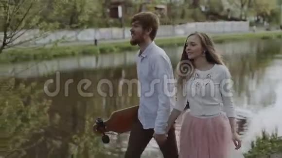 恋爱中的年轻夫妇牵着手在夏季公园散步穿着粉红色图塔裙的女人手里拿着长板视频的预览图