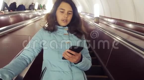 少女在地下地铁乘坐自动扶梯手持智能手机小女孩棕发女儿搜索视频的预览图