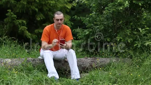 一个戴着红色智能手机的人坐在森林里的一棵倒下的树上视频的预览图
