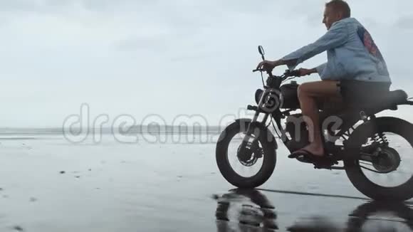 年轻英俊的嬉皮士骑着现代定制摩托车赛车在黑沙滩附近的水冲浪场地视频的预览图