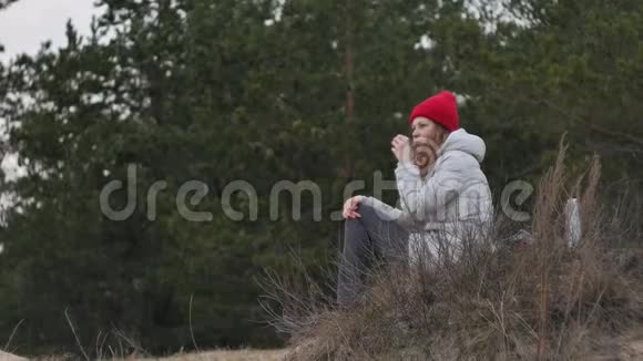 早春一位戴着红帽子的迷人年轻女子背着一个大旅游背包在森林里散步喝酒视频的预览图