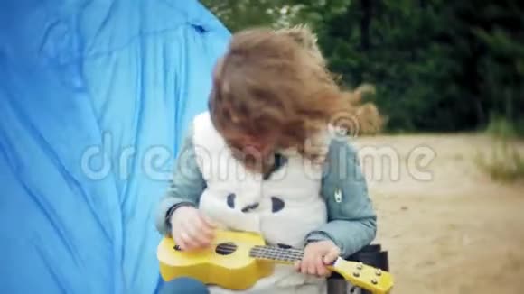 一个美丽的女孩和她的家人在一个旅游帐篷附近的河岸上的四弦琴上玩耍视频的预览图