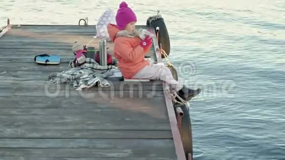 小女孩穿着暖和的衣服坐在河岸上坐在河港上喝热茶野餐视频的预览图