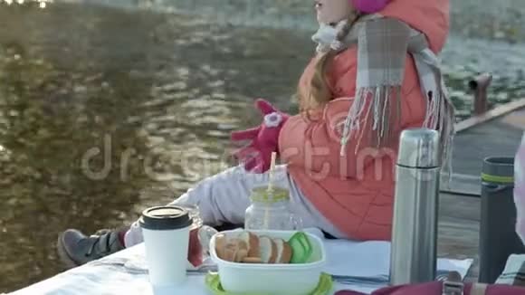小女孩穿着暖和的衣服坐在河岸上坐在河港上喝热茶野餐视频的预览图