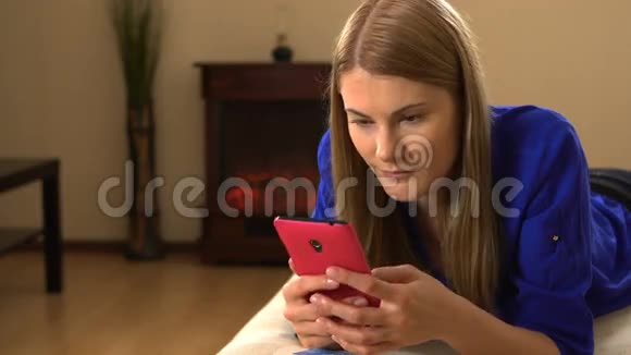 漂亮的年轻女人一个智能手机躺在沙发上浏览互联网和微笑视频的预览图
