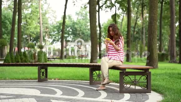 漂亮的女人坐在公园的长凳上拿着智能手机视频的预览图