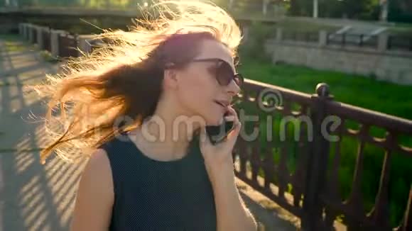 戴墨镜的女人在智能手机上说话在日落时分走在街上特写镜头视频的预览图