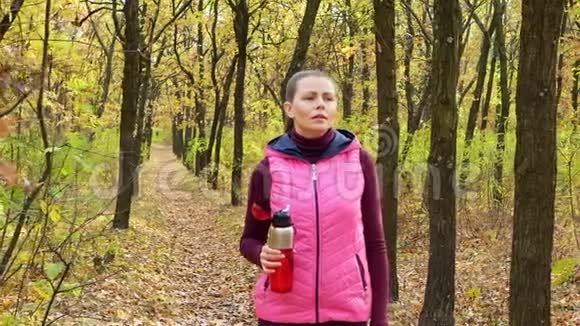 美丽的健身运动女孩走在秋季公园的运动服与运动水瓶或等渗饮料在手视频的预览图