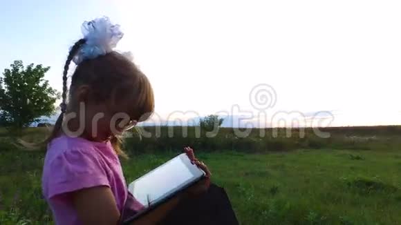 小女孩热情地在平板电脑上玩戴眼镜的女孩子站在绿色的草地上视频的预览图