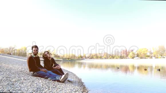 可爱的情侣坐在湖边带着耳机听音乐视频的预览图