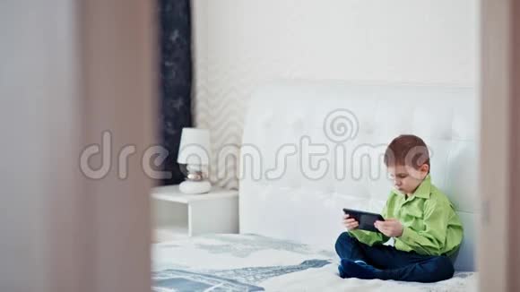 穿绿色衬衫的孩子正坐在床上玩平板电脑视频的预览图