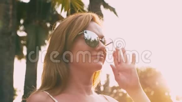 美丽时尚的金发女人戴着太阳镜沿着棕榈树的小径散步手掌倒映在眼镜上4K视频的预览图