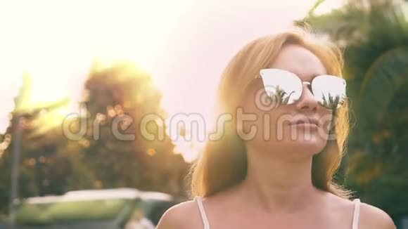 美丽时尚的金发女人戴着太阳镜沿着棕榈树的小径散步手掌倒映在眼镜上4K视频的预览图