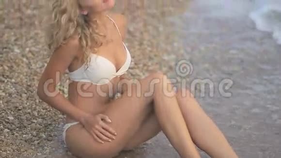 年轻漂亮的晒黑女孩穿着比基尼金发坐在海边的日落视频的预览图