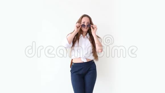 现代青年的时尚穿着牛仔裤白衬衫带皮背包和眼镜的时髦女孩在白色墙上摆姿势视频的预览图