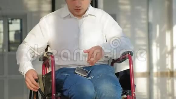 坐在轮椅上的残疾人在窗户旁听着智能手机耳机里的音乐视频的预览图