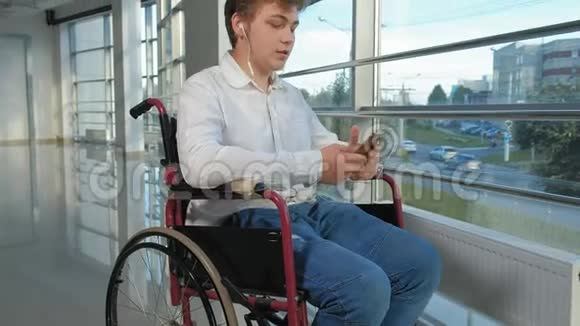 坐在轮椅上的残疾人在窗户旁听着智能手机耳机里的音乐视频的预览图