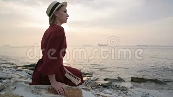 日落时分一个漂亮的白人女孩穿着一件红色鲜艳的连衣裙和一顶草帽坐在海边的一块大石头上看书视频的预览图