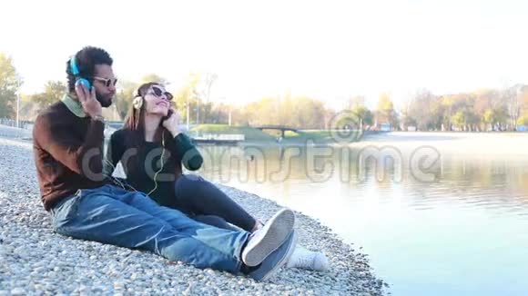 可爱的情侣坐在湖边带着耳机听音乐视频的预览图