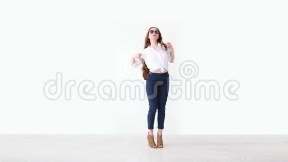 现代青年的时尚穿着牛仔裤白衬衫带皮背包和眼镜的时髦女孩在白色墙上摆姿势视频的预览图