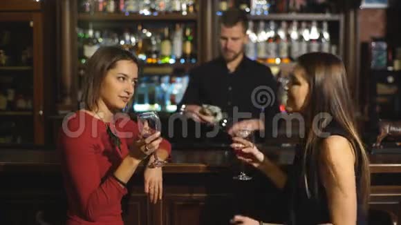 两个漂亮的年轻女人在酒吧里聚在一起喝一杯红酒坐在柜台前互相微笑视频的预览图
