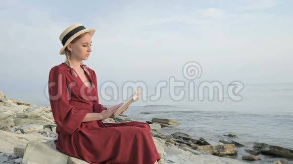 日落时分一个漂亮的白人女孩穿着一件红色鲜艳的连衣裙和一顶草帽坐在海边的一块大石头上看书视频的预览图