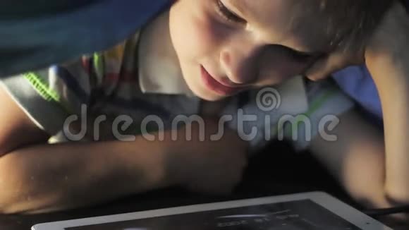男孩躺在毯子下在触摸板上看卡通视频的预览图