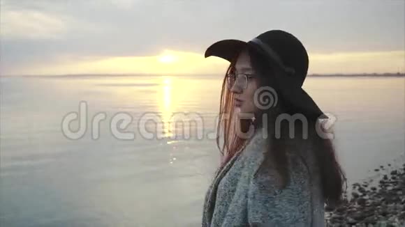 日落时分海边戴着黑色帽子和眼镜的年轻漂亮微笑的女人视频的预览图