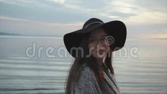 日落时分海边戴着黑色帽子和眼镜的年轻漂亮微笑的女人视频的预览图