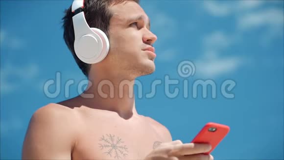英俊的年轻人用无线耳机听他的智能手机里的音乐在海滩上跳舞背景是蓝色视频的预览图