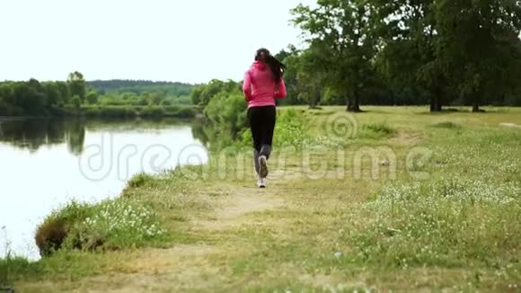 一个穿着粉红色夹克和黑色裤子的女孩戴着耳机在河边跑步为马拉松做准备视频的预览图