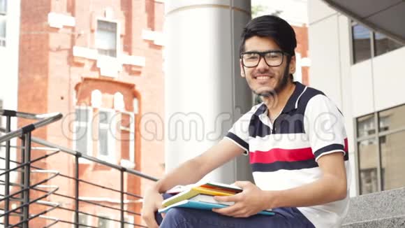 一位微笑的亚裔男学生坐在楼梯上看书的画像视频的预览图