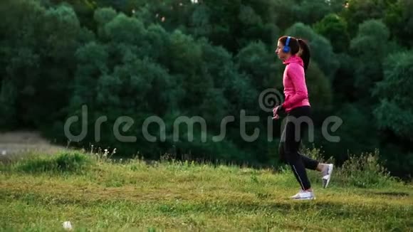 一个穿着粉红色夹克和黑色裤子的女孩戴着耳机在河边跑步为马拉松做准备视频的预览图